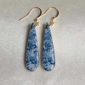 May include: A pair of gold dangle earrings with blue and white floral patterns. The earrings are teardrop shaped and have a delicate design.
