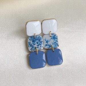 May include: A pair of blue and white ceramic earrings with a floral pattern. The earrings are square shaped and have gold accents.