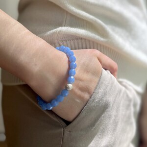 May include: A light blue beaded bracelet with a silver charm. The bracelet is made of faceted beads and is worn on a person's wrist.