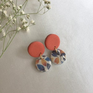 Könnte beinhalten: Ein Paar korallenfarbene Ohrringe aus Ton mit einem weißen und blauen Blumenmuster. Die Ohrringe sind rund und haben einen goldenen Akzent.