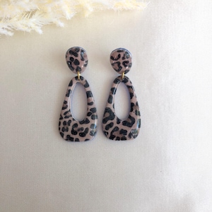 May include: A pair of teardrop-shaped earrings with a leopard print design. The earrings have a light brown base color with black spots. The top of each earring is a small, round stud. The earrings are displayed on a white surface.