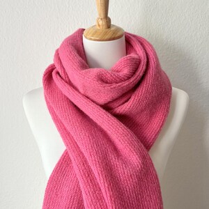 May include: A vibrant pink knitted scarf draped over a white mannequin. The scarf is made of a thick, textured yarn, and is displayed against a plain white background. The scarf is a solid color, with no visible patterns or embellishments.