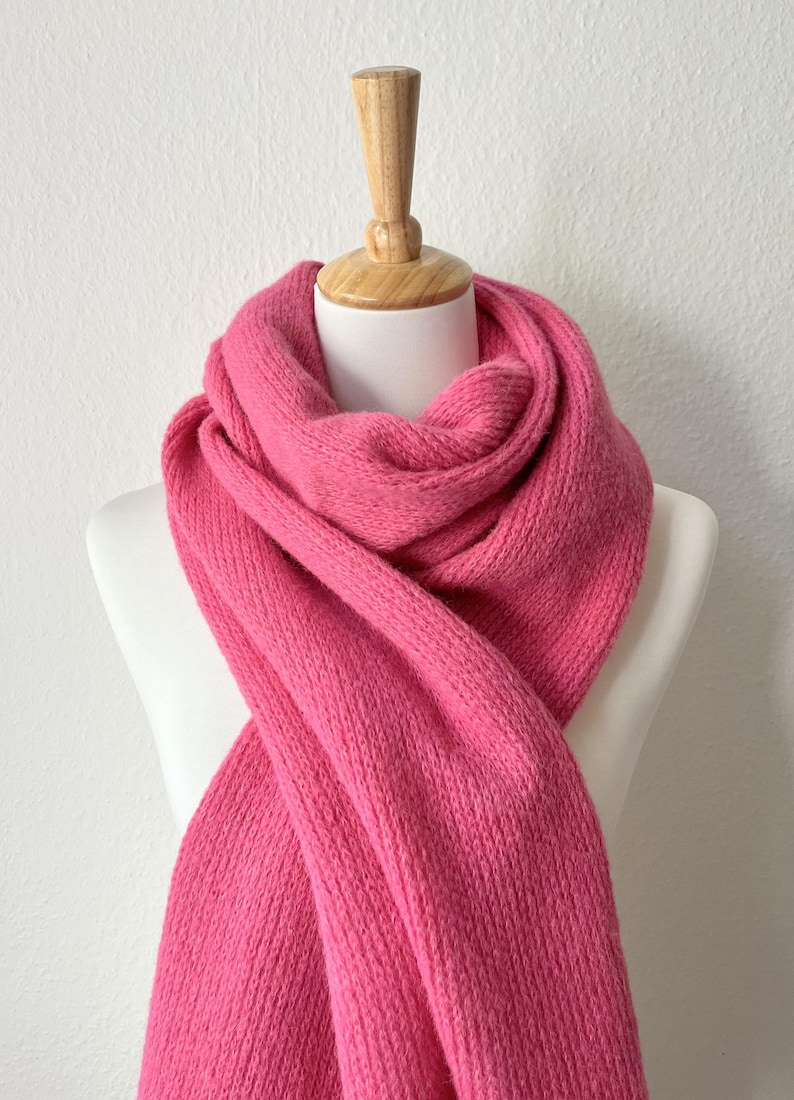 May include: A vibrant pink knitted scarf draped over a white mannequin. The scarf is made of a thick, textured yarn, and is displayed against a plain white background. The scarf is a solid color, with no visible patterns or embellishments.