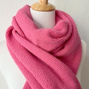 May include: A vibrant pink scarf, draped over a white mannequin head. The scarf is made of a soft, knitted material. The bright color and texture of the scarf are the focal points of the image.