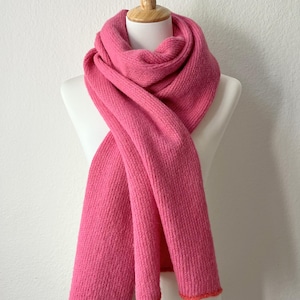 May include: A vibrant pink scarf displayed on a mannequin. The scarf is made of a thick, knitted material and has a contrasting orange trim along the edges. The scarf is draped in a way that showcases its length and texture.