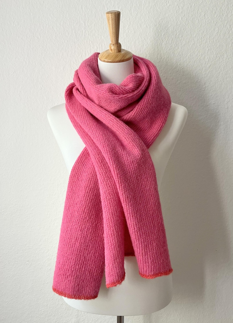 May include: A vibrant pink scarf displayed on a mannequin. The scarf is made of a thick, knitted material and has a contrasting orange trim along the edges. The scarf is draped in a way that showcases its length and texture.