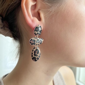 Peut inclure: Boucles d'oreilles pendantes à trois niveaux, de forme ovale et à motif léopard. Les boucles d'oreilles sont faites d'un matériau de couleur claire avec des taches noires et marron. Les boucles d'oreilles sont suspendues à l'oreille d'une personne.