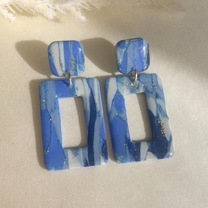 Könnte beinhalten: Ein Paar handgefertigte Ohrringe aus Polymer Clay. Jeder Ohrring hat einen quadratischen oberen Teil und einen rechteckigen Tropfen mit einem Ausschnitt. Die Ohrringe sind mit Blautönen und Weiß marmoriert und mit Goldflocken akzentuiert.