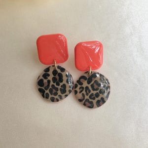Könnte beinhalten: Ein Paar Ohrhänger. Jeder Ohrring hat einen glänzenden, quadratischen, orangefarbenen oberen Teil und einen runden, leopardenmusterartigen unteren Teil. Die Ohrringe sind durch eine goldfarbene Metallschlaufe verbunden. Der Hintergrund ist ein hellbeiger Stoff.