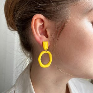 Könnte beinhalten: Hellgelbe Ohrringe mit einem rechteckigen Stecker und einem strukturierten, unregelmäßigen Kreisanhänger. Die Ohrringe sind ein auffälliges, modernes Accessoire. Ein auffälliges Schmuckstück.