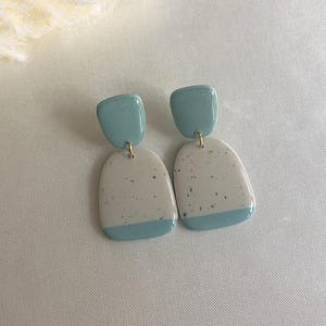 Peut inclure: Une paire de boucles d'oreilles faites à la main. Chaque boucle d'oreille présente un dessus carré bleu clair relié à un bas rectangulaire blanc moucheté et bleu clair. Les boucles d'oreilles sont reliées par une petite boucle dorée.