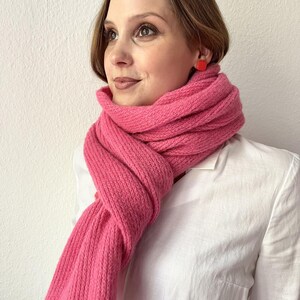 May include: A vibrant pink knitted scarf with a contrasting red trim. The scarf is wrapped around the neck and shoulders, creating a cozy and stylish look. The scarf is paired with a white collared shirt and red square earrings.