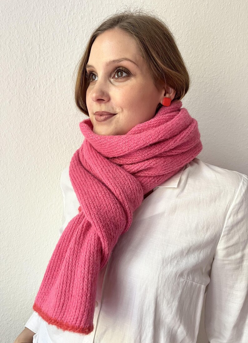 May include: A vibrant pink knitted scarf with a contrasting red trim. The scarf is wrapped around the neck and shoulders, creating a cozy and stylish look. The scarf is paired with a white collared shirt and red square earrings.