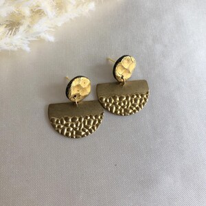 Puede incluir: Un par de pendientes dorados. Cada pendiente presenta una mitad inferior semicircular texturizada y una parte superior circular más pequeña. Los pendientes están colocados sobre un fondo neutro, con un toque de flores secas.