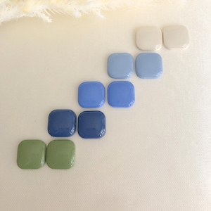 Könnte beinhalten: Eine Sammlung quadratischer Glasfliesen in verschiedenen Blau-, Grün- und Cremetönen. Die Fliesen sind diagonal angeordnet und zeigen ihre glänzende Oberfläche und Farbvariationen. Der Hintergrund ist ein weicher, cremefarbener Stoff.