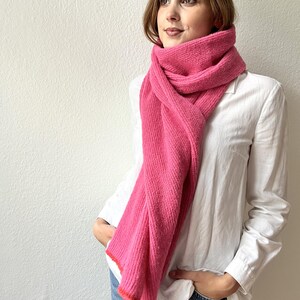 May include: A bright pink scarf with a ribbed texture is draped around the neck. The scarf has a contrasting orange trim. The person is wearing a white button-down shirt and blue jeans.