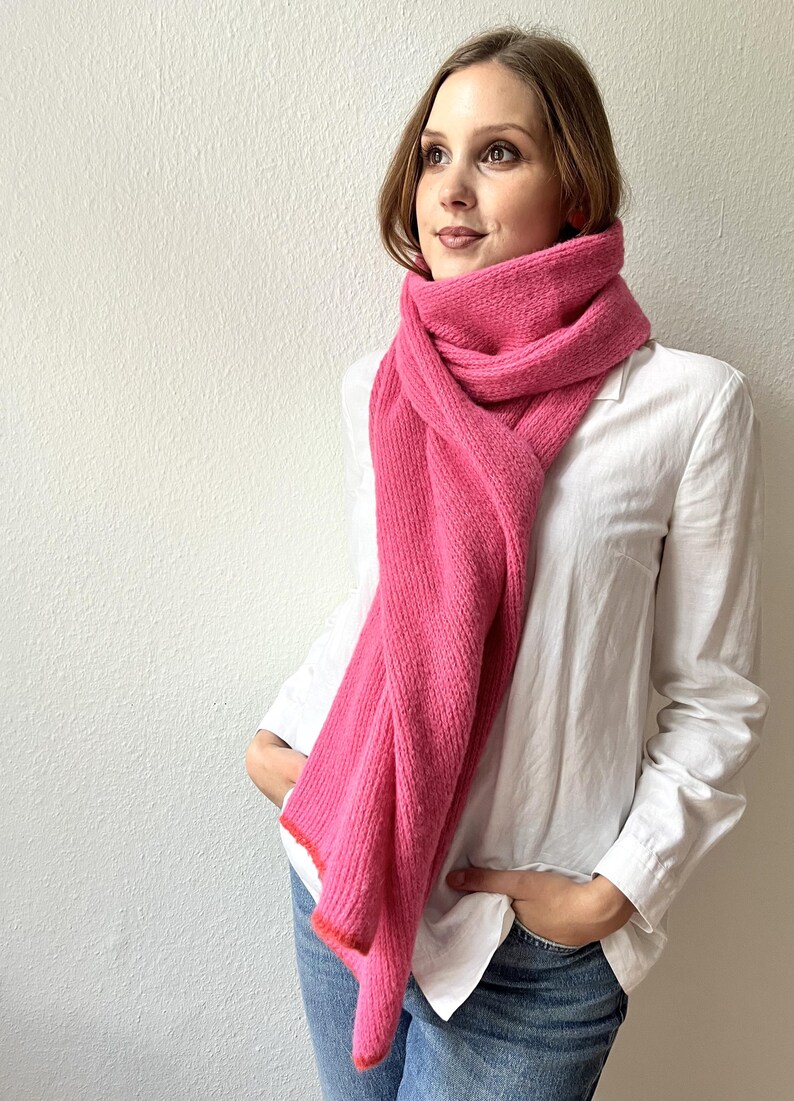 May include: A bright pink scarf with a ribbed texture is draped around the neck. The scarf has a contrasting orange trim. The person is wearing a white button-down shirt and blue jeans.