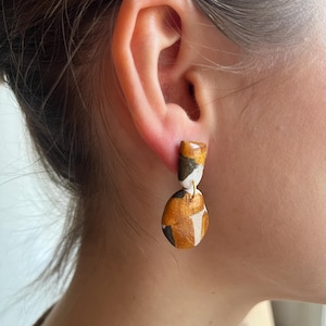 Op de afbeelding: Een paar abstracte oorhangers. De oorbellen hebben een rechthoekige bovenkant en een druppelvormige onderkant, verbonden door een goudkleurige draad. De oorbellen hebben een patroon van witte, zwarte en gouden geometrische vormen.