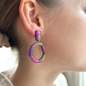May include: A pair of colourful earrings. The earrings feature a rectangular top in shades of purple and green, connected to a rounded, open-circle design in similar hues. The earrings are a unique accessory.