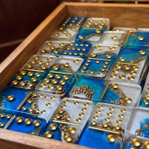May include: A set of dominoes in a wooden box. The dominoes have a clear resin base with blue and white accents, and gold-colored dots. The box is open, revealing the dominoes inside.