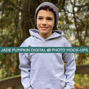 May include: A young person wearing a gray hooded sweatshirt and a gray beanie. The sweatshirt has a front pocket and is paired with blue jeans. The text "JADE PUMPKIN DIGITAL PHOTO MOCK-UPS" is displayed across the bottom of the image.