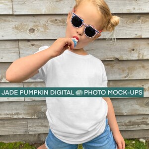 May include: A young girl wearing a white t-shirt and blue denim shorts with a white lace trim. She is wearing red, white and blue striped sunglasses and is holding a blue and white popsicle.