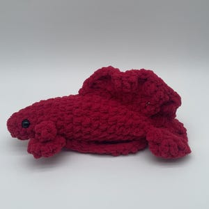May include: A handmade, plush, red crocheted lobster toy. The lobster has a textured, chunky appearance with a rounded body, small claws, and a tail. It features a single black eye and is set against a white background.