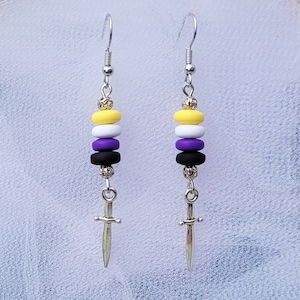 May include: A pair of silver earrings with a sword charm and four beads in the colours of the non-binary pride flag: yellow, white, purple, and black.