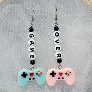 Puede incluir: Pendientes colgantes con temática de videojuegos. Un pendiente presenta la palabra "GAME" deletreada en cuentas de letras blancas, el otro dice "OVER". Cada pendiente tiene un dije de controlador en azul claro y rosa.