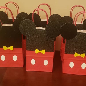 K&ouml;nnte beinhalten: Rote und schwarze Geschenkt&uuml;ten im Mickey-Mouse-Design mit gelben Schleifen und wei&szlig;en Punkten. Die Taschen haben Glitzer auf den schwarzen Ohren und dem Gesicht.