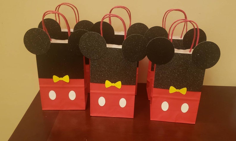 K&ouml;nnte beinhalten: Rote und schwarze Geschenkt&uuml;ten im Mickey-Mouse-Design mit gelben Schleifen und wei&szlig;en Punkten. Die Taschen haben Glitzer auf den schwarzen Ohren und dem Gesicht.
