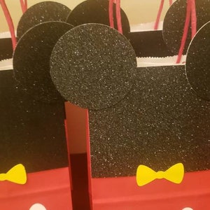 K&ouml;nnte beinhalten: Ein Set mit f&uuml;nf roten und schwarzen Geschenkt&uuml;ten im Mickey-Mouse-Design mit gelben Schleifen. Die Taschen sind aus Papier und haben Glitzer auf den schwarzen Teilen.