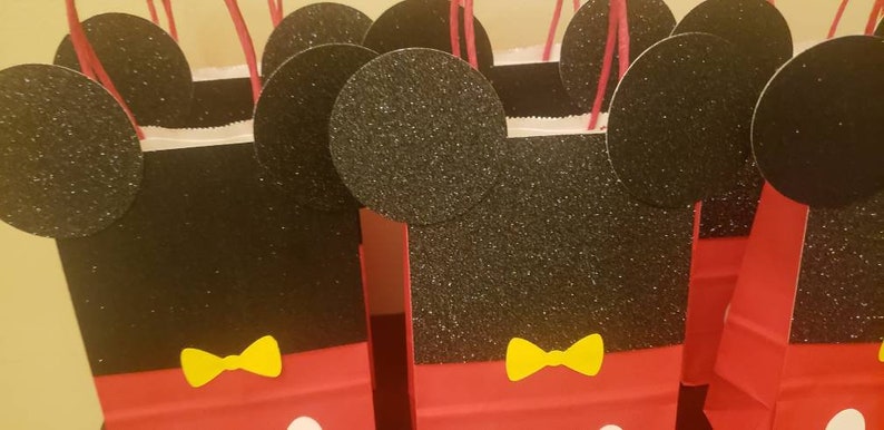 K&ouml;nnte beinhalten: Ein Set mit f&uuml;nf roten und schwarzen Geschenkt&uuml;ten im Mickey-Mouse-Design mit gelben Schleifen. Die Taschen sind aus Papier und haben Glitzer auf den schwarzen Teilen.
