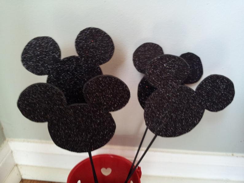 Puede incluir: Cinco orejas de Mickey Mouse de fieltro negro con purpurina, dispuestas en un racimo. Las orejas est&aacute;n unidas a palos y est&aacute;n en un cubo rojo.