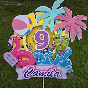 May include: A colorful cake topper with a beach theme. It features a pink flamingo with an ice pop, a beach ball, palm trees, and the number 9. The name "Camila" is written on a banner, with a background of water splashes and confetti.