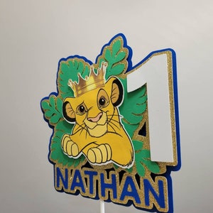 Op de afbeelding: Een gelaagde taarttopper met een cartoon leeuw met een kroon, het nummer 1 en de naam "NATHAN". Het ontwerp bevat groene, gouden, blauwe en witte elementen, met glitteraccenten. Voor een verjaardagsfeest.