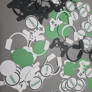 May include: Black and green confetti with the name "Daniel" printed on white circles. The confetti includes images of video game controllers and headphones.