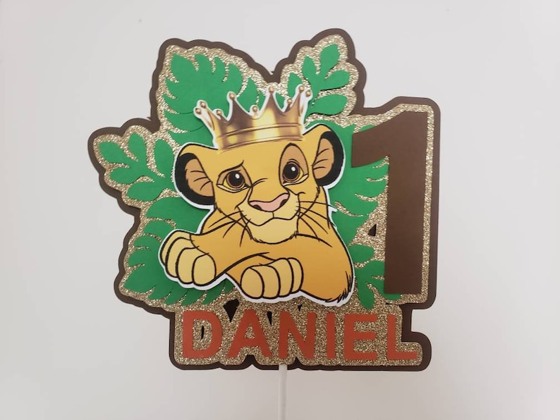 Op de afbeelding: Een taarttopper met Simba uit The Lion King, met een gouden kroon, het nummer 1 en de naam "DANIEL" in oranje. Het ontwerp bevat groene bladeren en gouden glitteraccenten, geschikt voor een eerste verjaardag.