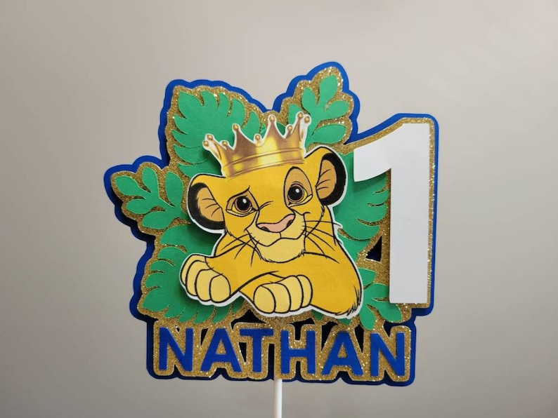 Op de afbeelding: Een gepersonaliseerde taarttopper met een cartoon leeuw met een kroon, het nummer 1 en de naam "NATHAN". Het ontwerp bevat blauwe, groene, goudglitter en witte elementen, perfect voor een eerste verjaardag.