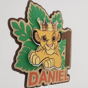 Op de afbeelding: Een gelaagde taarttopper met een cartoon leeuwenwelp met een gouden kroon, het nummer 1 en de naam "DANIEL" in oranje letters. Het ontwerp bevat groene bladeren en gouden glitteraccenten, perfect voor een verjaardag.