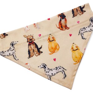 Puede incluir: Un pañuelo beige para perro con un patrón repetido de varias razas de perros en estilo acuarela. El pañuelo presenta corazones rosas y una variedad de razas de perros, incluyendo un dálmata, un terrier y un golden retriever.