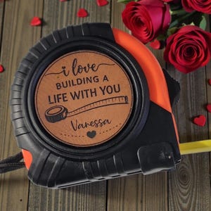 May include: Black and orange tape measure with a brown leather-like center. The center reads "i love BUILDING A LIFE WITH YOU" with the name "Vanessa" and a heart. Red roses and heart-shaped confetti are in the background.