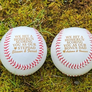 May include: Two white baseballs with red stitching. The baseballs have gold text that reads "We hit a homerun scoring you as our stepdad Eleanor & Farrah" and "We hit a homerun scoring you as our bonus dad Autumn & Vanessa".