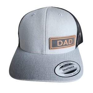 May include: Gray and black trucker hat with a curved brim. The hat features a rectangular patch with the word "DAD" in a copper color. A white and black circular tag is on the brim.