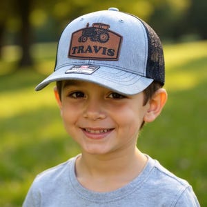 May include: A light blue and black trucker hat with a brown patch featuring a tractor graphic and the name "TRAVIS". The hat is worn by a smiling child, with a gray t-shirt.