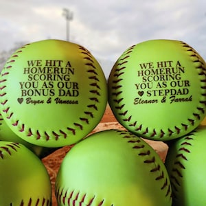 May include: Two green softball-shaped gifts with personalized messages. The first softball says "We hit a homerun scoring you as our bonus dad Bryan & Vanessa". The second softball says "We hit a homerun scoring you as our stepdad Eleanor & Farrah".