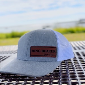 May include: A gray and light blue trucker hat with a brown leather patch that says "RING BEARER" with a signature underneath.