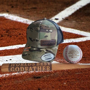 May include: A camouflage baseball cap with "GODFATHER" patch, a baseball with "I HIT A HOMERUN SCORING YOU AS MY GODFATHER" text, and a wooden bat with "WORLDS BEST GODFATHER" engraved. The items are on a baseball field.