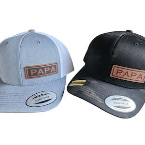 May include: Two trucker hats, one gray and one black, each with a "PAPA" patch. The gray hat has a white mesh back, while the black hat has a camouflage pattern. Both have a snapback closure.