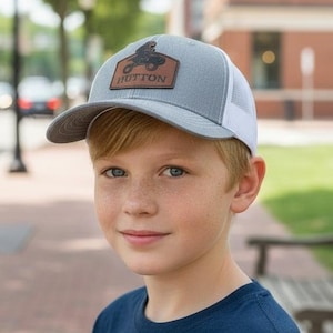 May include: Gray trucker hat with a white mesh back and a curved brim. The hat features a brown leather patch with a motorcycle graphic and the name "HUTTON". The hat is worn by a person.